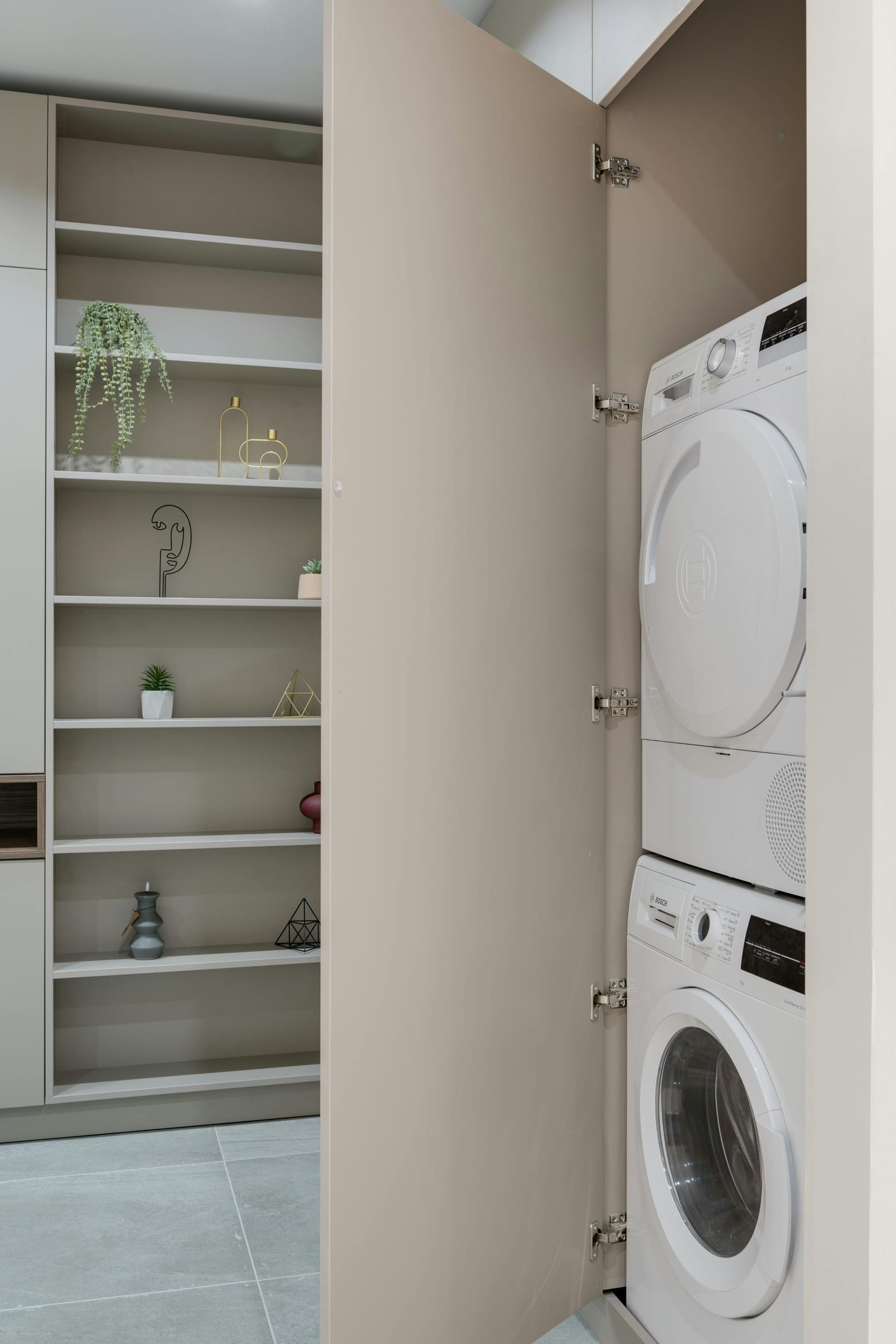 Stacked Bosch dryer and washing machine concealed inside a cabinet, with the cabinet door open beside a neutral shelving unit