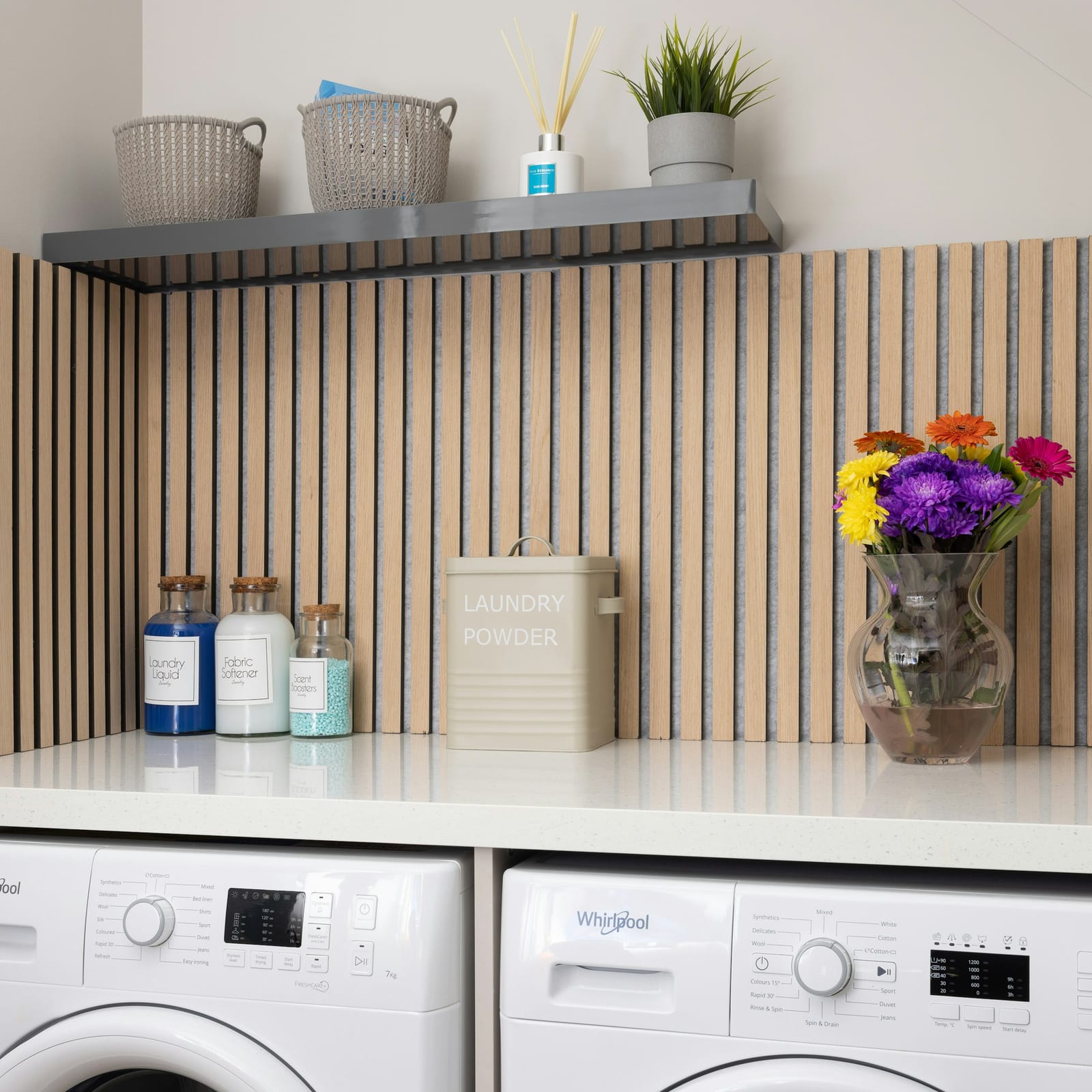 Modern Whirlpool front-loader washing machine and dryer side by side under a fluted wood backsplash with a laundry powder tin and flowers on the bench