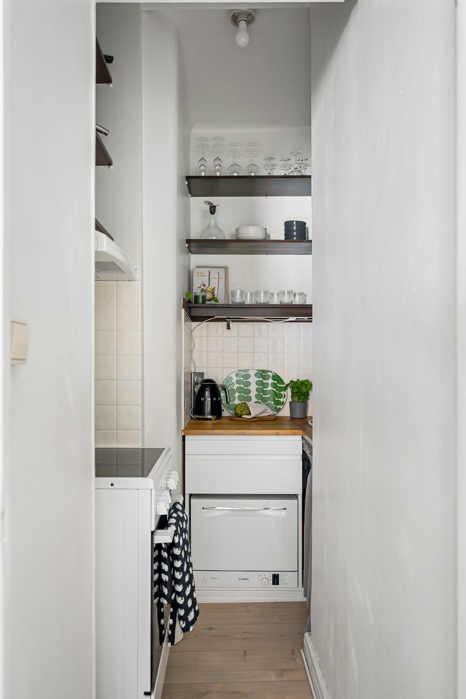 Compact rental apartment kitchen with white cabinetry, a SMEG kettle on a timber benchtop, built-in oven, cooktop and Bosch dishwasher visible through the doorway
